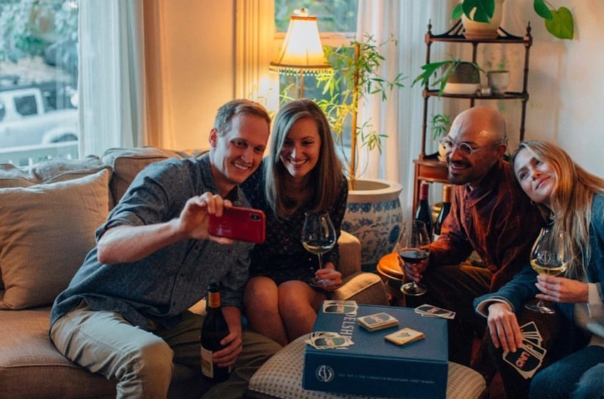 Four adults smiling indoors while a man takes a photo with his phone, with a board game and wine bottles on the table in front of them.