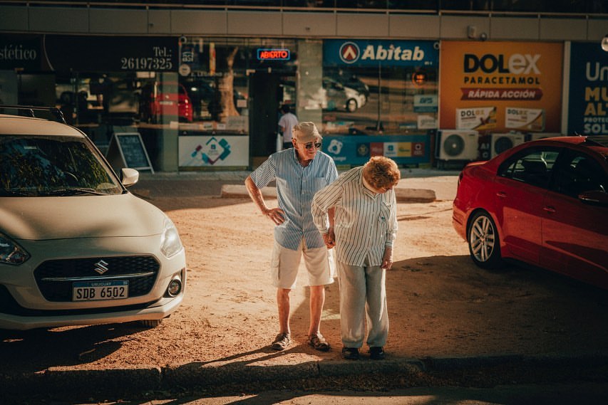 Elderly couple walking hand-in-hand on a sunny street near parked cars. Woman leans slightly, man stands upright in casual clothing.