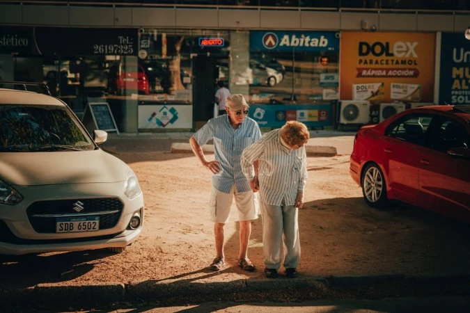 Elderly couple walking hand-in-hand on a sunny street near parked cars. Woman leans slightly, man stands upright in casual clothing.