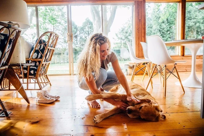 A smiling adult woman pets a golden retriever dog indoors on a wooden floor, with bright daylight streaming through a window in the background.