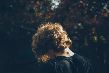 Close-up portrait of a person with short curly hair facing away, wearing a black t-shirt outdoors with bokeh foliage background