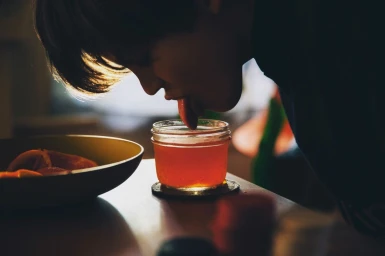 Close-up of a child leaning over a wooden table, sticking their tongue into a small glass jar filled with red jam.