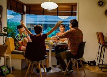 Family dining together in a cozy home interior with a child raising hands up at a table by a large window.