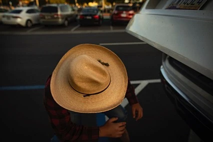Person wearing a straw cowboy hat and plaid shirt crouching near the open trunk of a white car in a parking lot