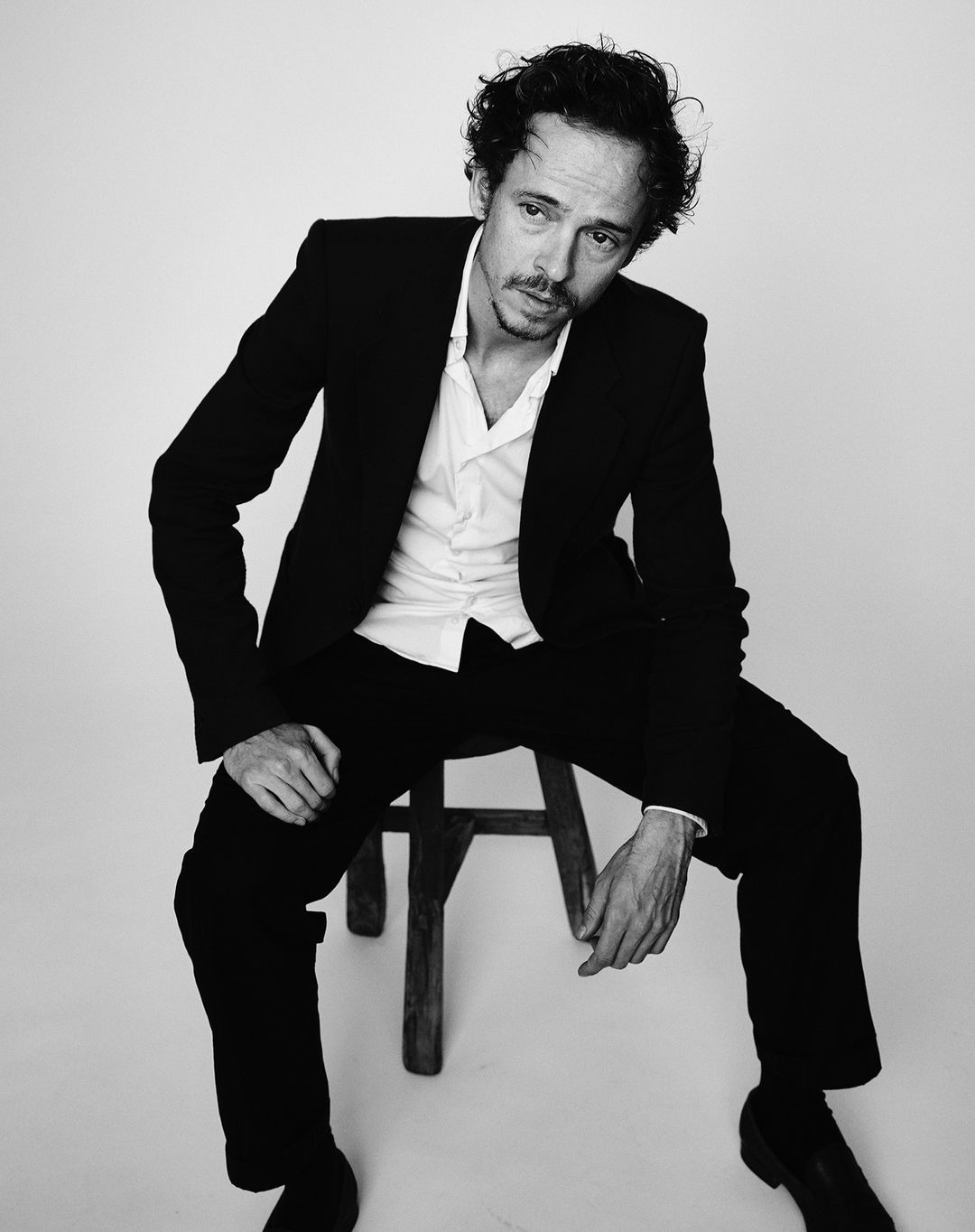 Black and white portrait of a man wearing a suit, seated on a stool in a photo studio with a neutral backdrop, looking off-camera.