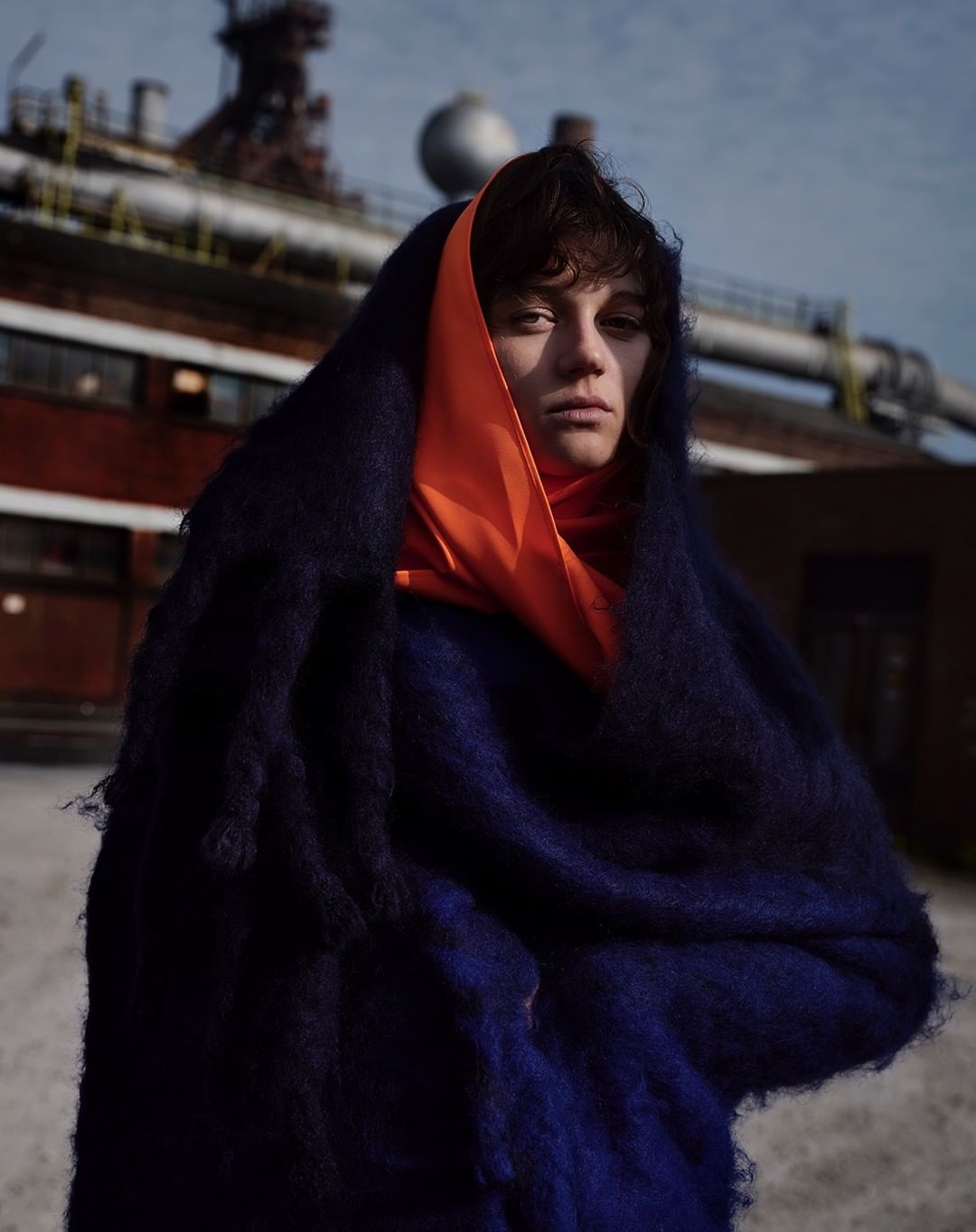 Woman wrapped in blue shawl with orange scarf, posing outdoors in front of an industrial factory under a slightly overcast sky.
