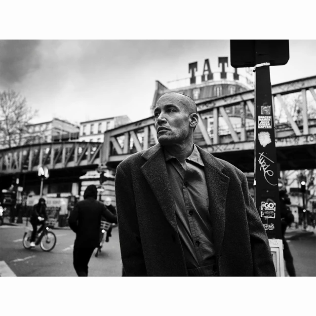 Black and white portrait of an adult man in a suit jacket looking to the side against a city background with a bridge.