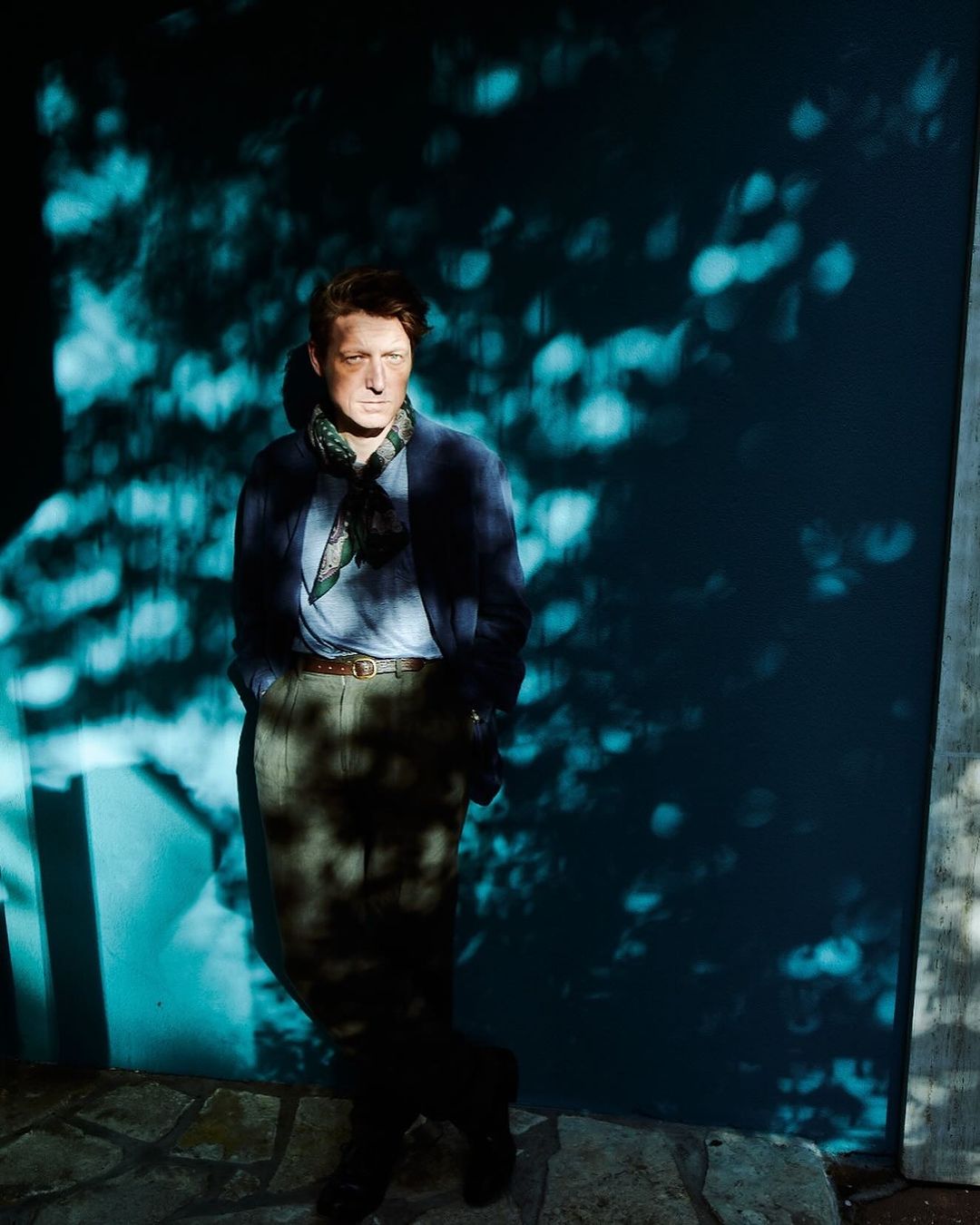 Portrait of a mature adult standing outdoors against an aqua-colored wall in dappled light, wearing a blazer and scarf.