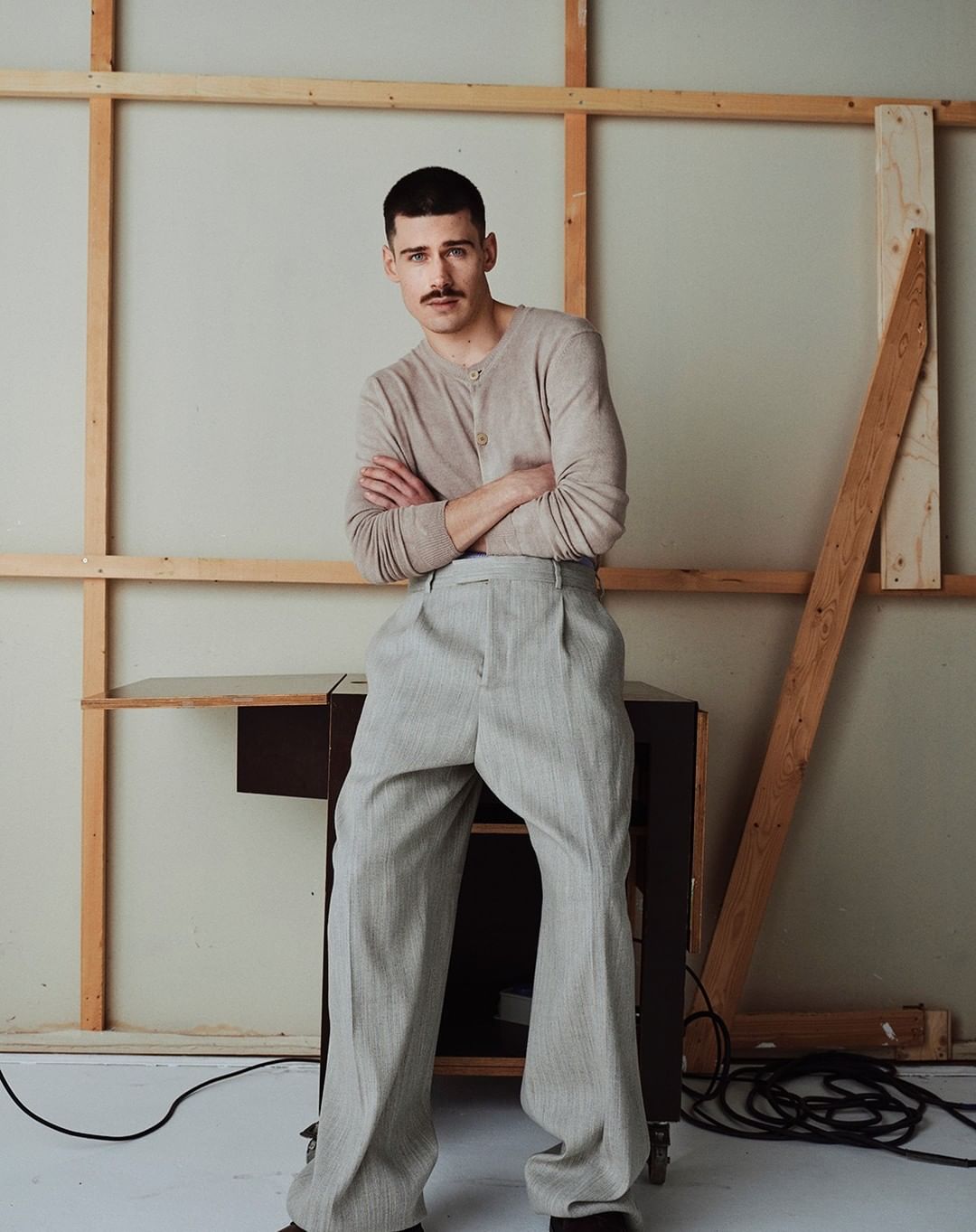Portrait of a young adult male with a mustache, wearing a long-sleeved shirt and high-waisted trousers, posing in front of a backdrop.