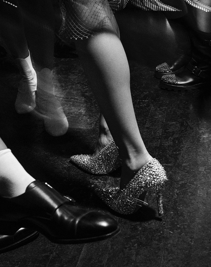 Monochrome shoe-level shot of woman in glittering high heels, alongside blurry motion of others wearing dark and white shoes.