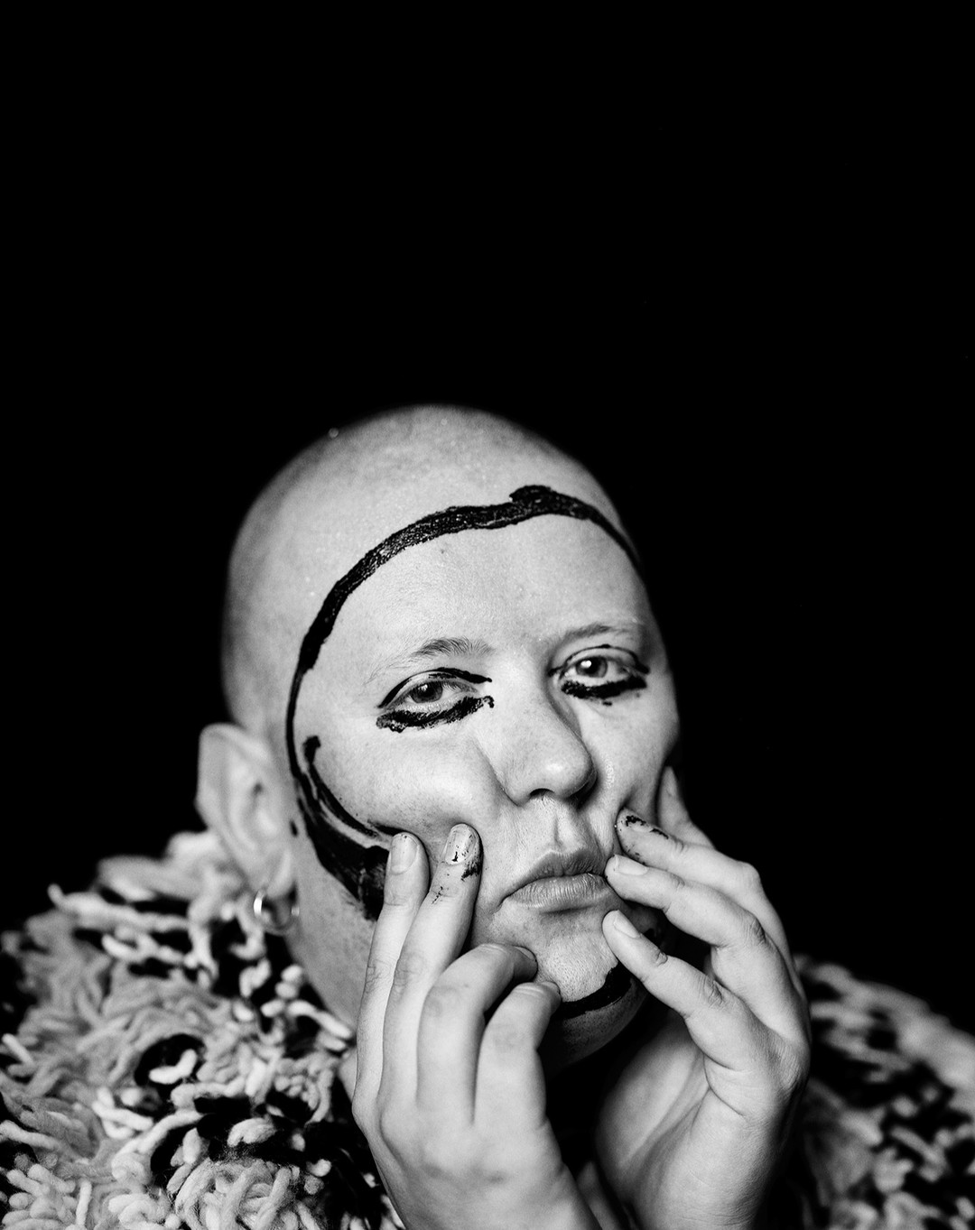 Dramatic black and white portrait of a bald person with painted face, expressive hands, and wearing textured clothing against black.