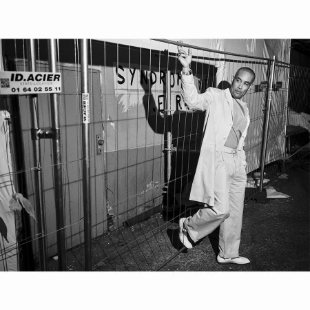 Man wearing a light suit and trench coat standing behind a metal construction fence in Paris by Charlie De Keersmaecker.