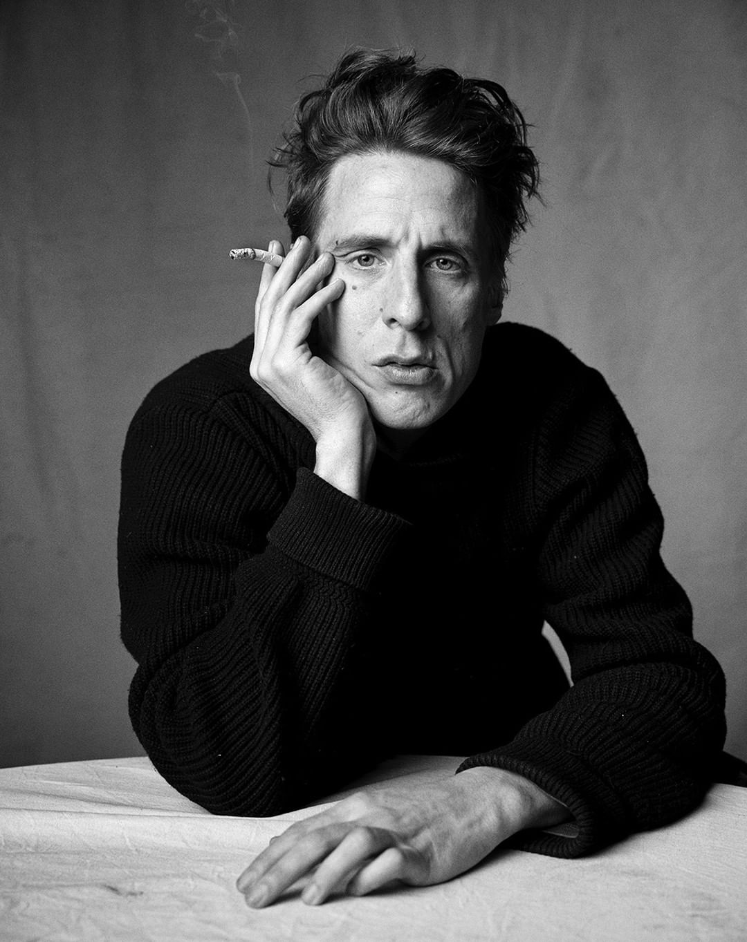 Black and white portrait of adult male with a cigarette in his mouth, resting his face on his hand and posed at a table.