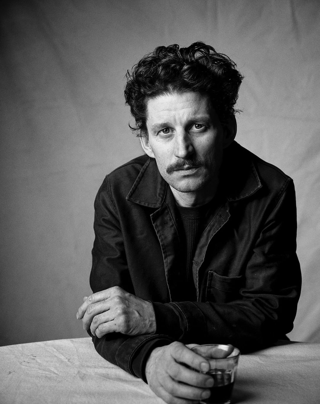 Monochrome portrait of adult male with dark curly hair and moustache, wearing a dark jacket, with a glass of liquid on a table.