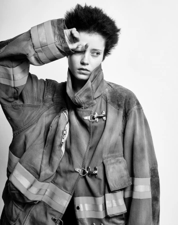 Monochrome portrait of a person in a firefighter jacket, hand shielding eyes against a bright white background in a studio setting.