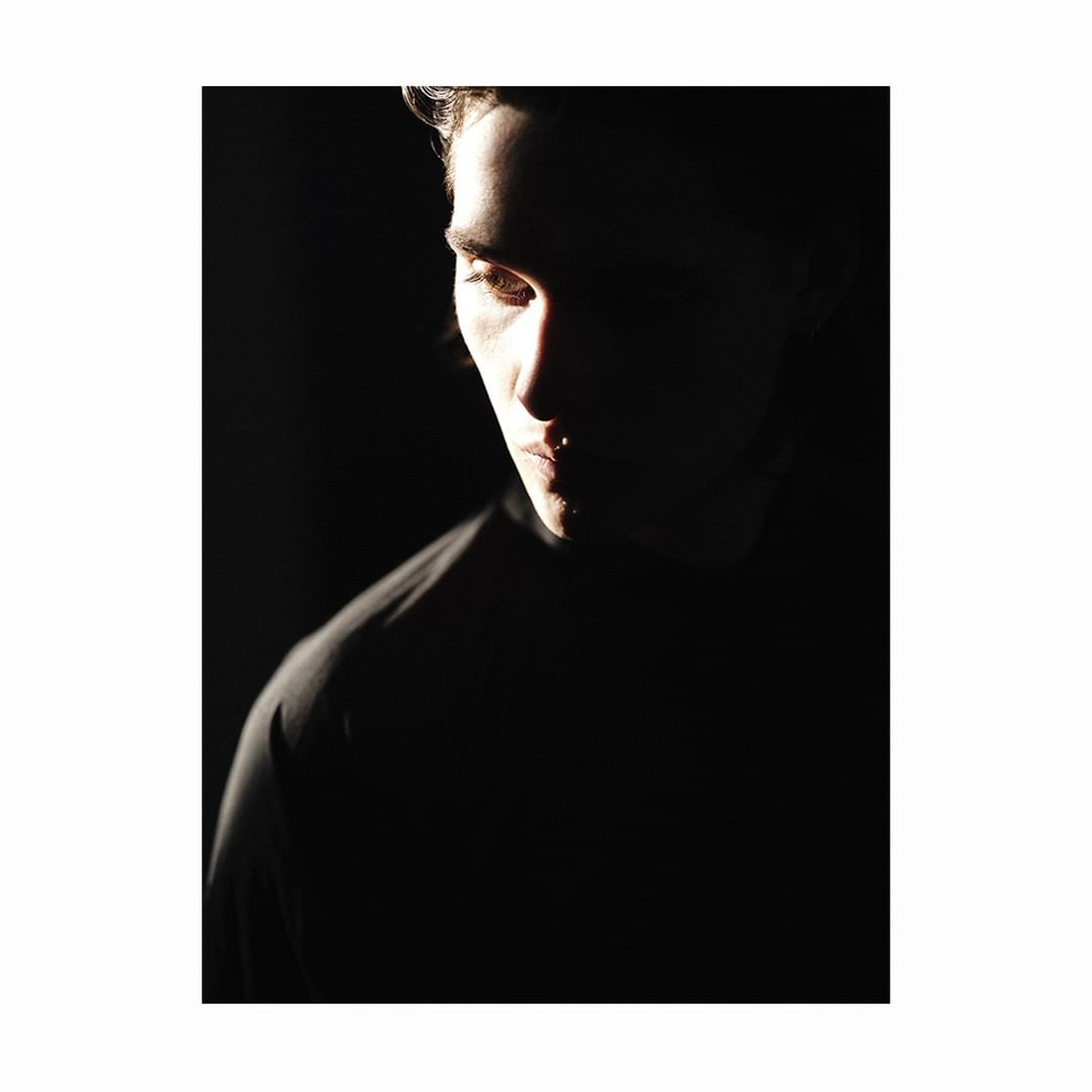 Dramatic portrait of a young adult male bathed in chiaroscuro lighting against a stark black backdrop, emphasizing facial features.