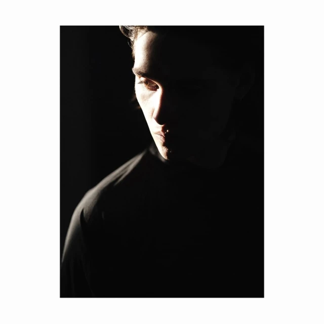 Dramatic portrait of a young adult male bathed in chiaroscuro lighting against a stark black backdrop, emphasizing facial features.