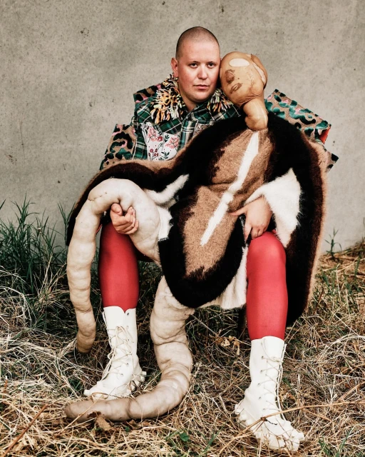 A fair-skinned person with a shaved head embraces a sculptural figure outdoors, wearing colorful clothing and red tights.