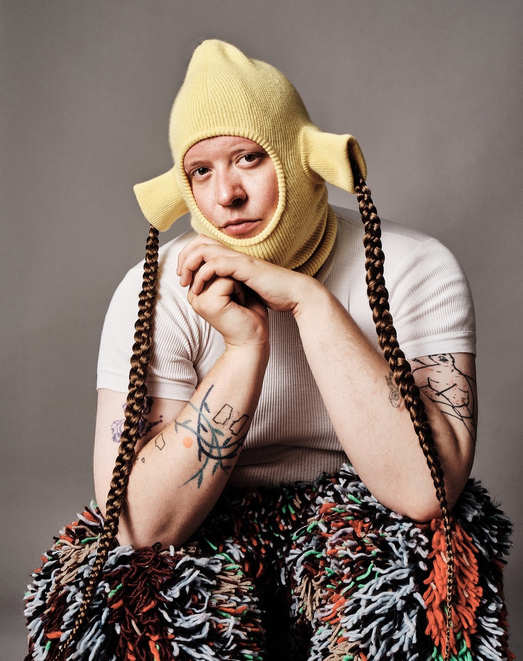 Person in yellow balaclava hat and fringed pants sits with hands clasped against a neutral gray background in a studio portrait.