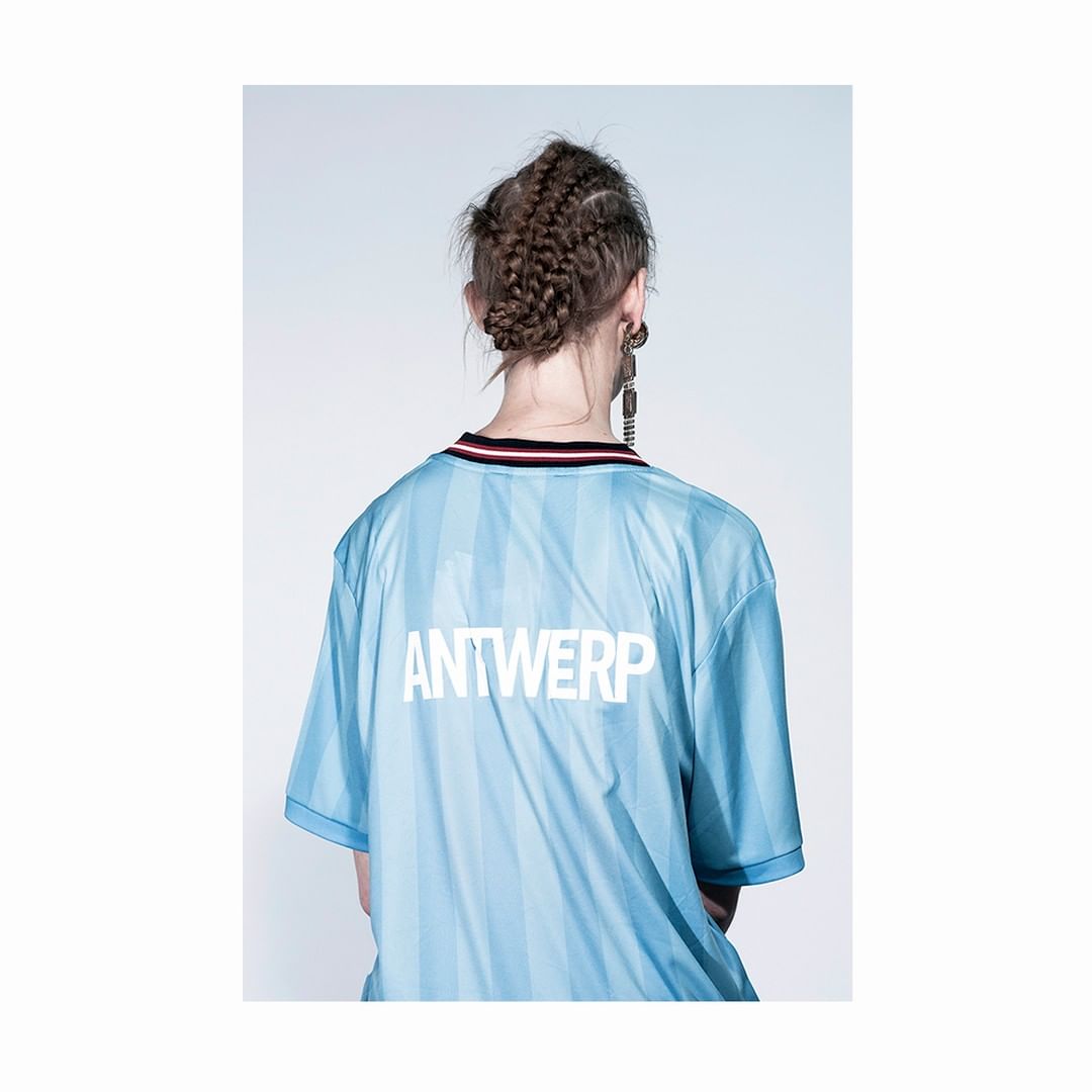 Back view of an adult female wearing a light blue jersey with 'ANTWERP' printed in white; she has braided hair and statement earrings.