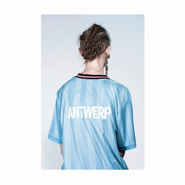 Back view of an adult female wearing a light blue jersey with 'ANTWERP' printed in white; she has braided hair and statement earrings.
