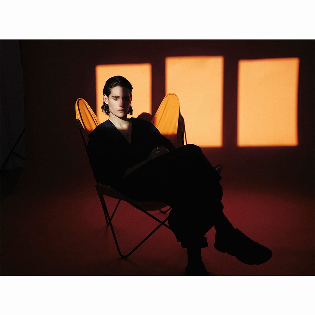 Adult male sits in a butterfly chair bathed in orange light with squared light patterns visible on the background wall.
