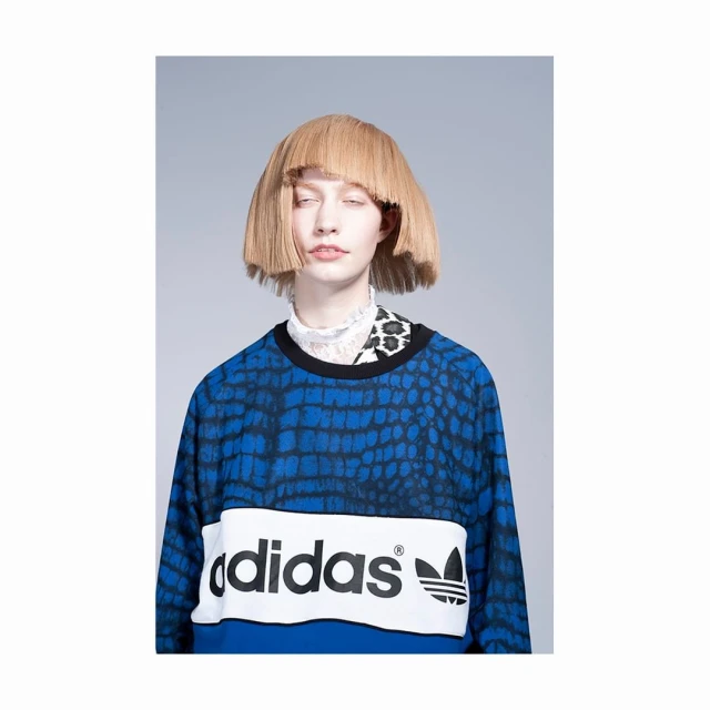 A woman with a blunt blonde wig poses in an Adidas sweatshirt featuring a blue animal print and the brand's logo centered on the garment.