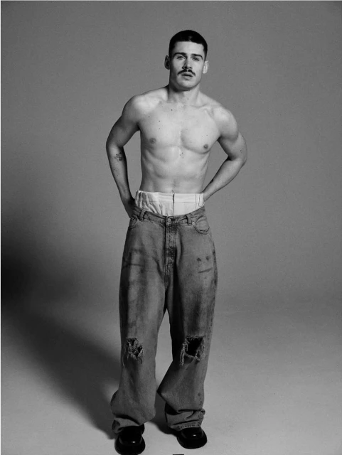 Full-length black and white studio portrait of a male model with a moustache, bare torso, ripped jeans and black shoes, hands on hips.