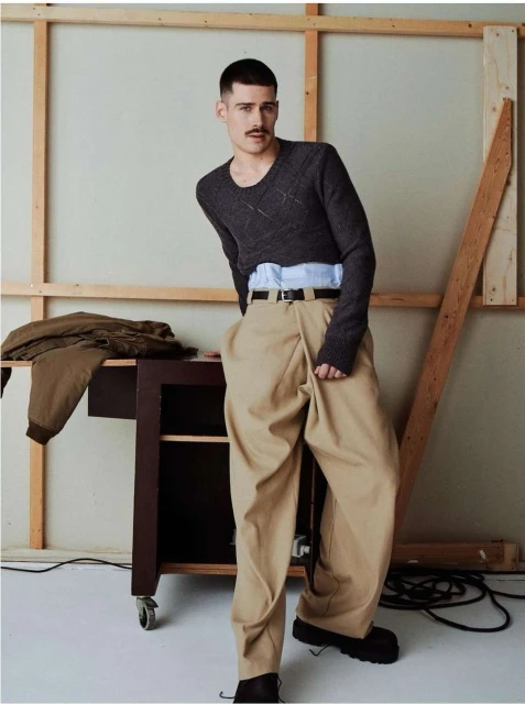 Male fashion model poses in a studio wearing a dark sweater, light blue dress shirt and tan oversized pants with black leather shoes.