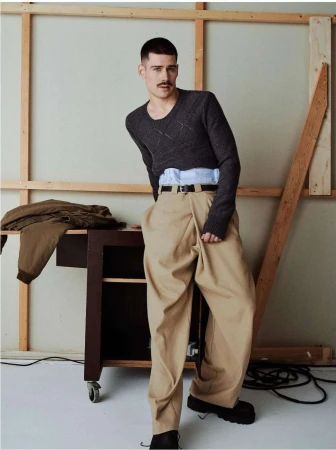Man with mustache wearing a grey sweater, blue shirt, and wide tan trousers leaning against a wooden studio frame