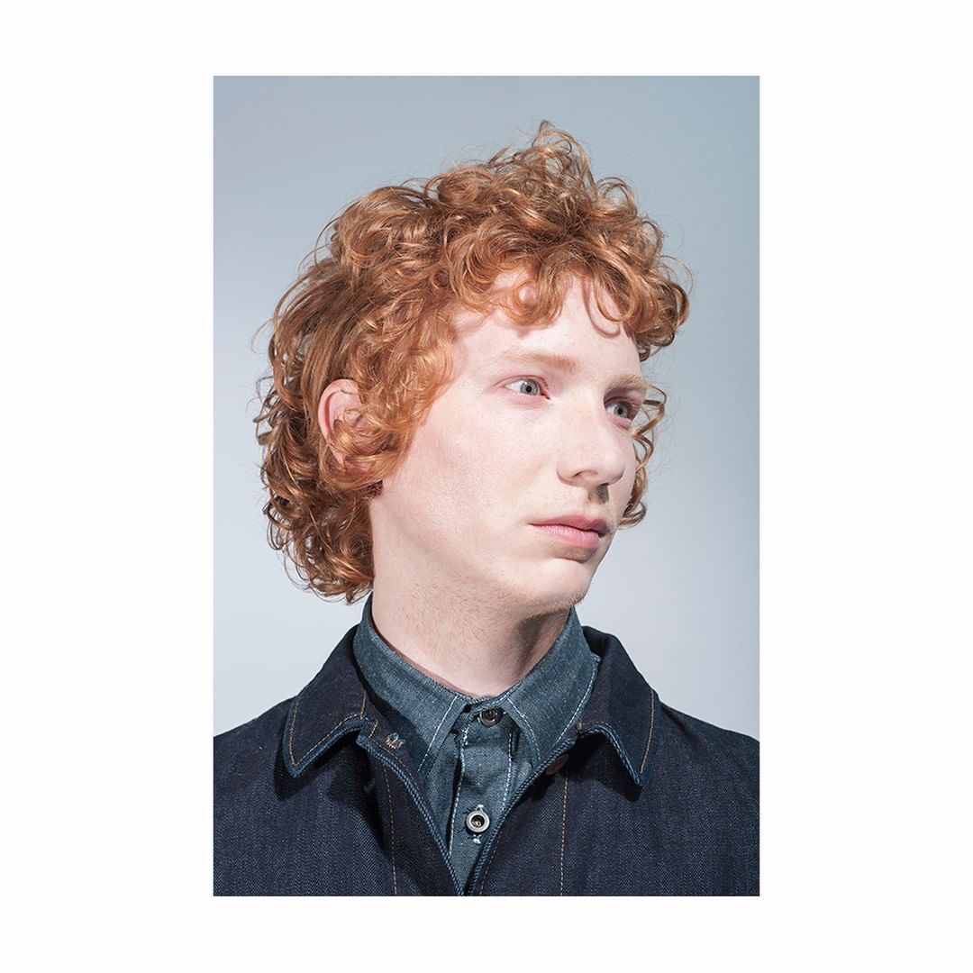 Close-up studio shot of a redhead male model with curly hair wearing a denim shirt and jacket. The background is light grey.