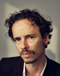 Close-up portrait of a man with curly brown hair and a mustache wearing a dark suit jacket and white shirt