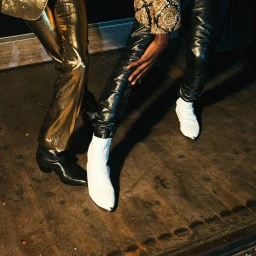 Close-up of two people's legs in metallic gold trousers and black leather pants standing on a wooden floor.