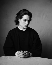 Monochrome portrait of a young man with dark hair wearing a dark crewneck sweater with clasped hands on a table.