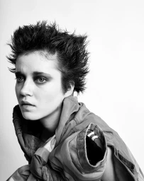 Black and white portrait of a woman with short spiky hair and dark eye makeup wearing a collared jacket against a plain white background.