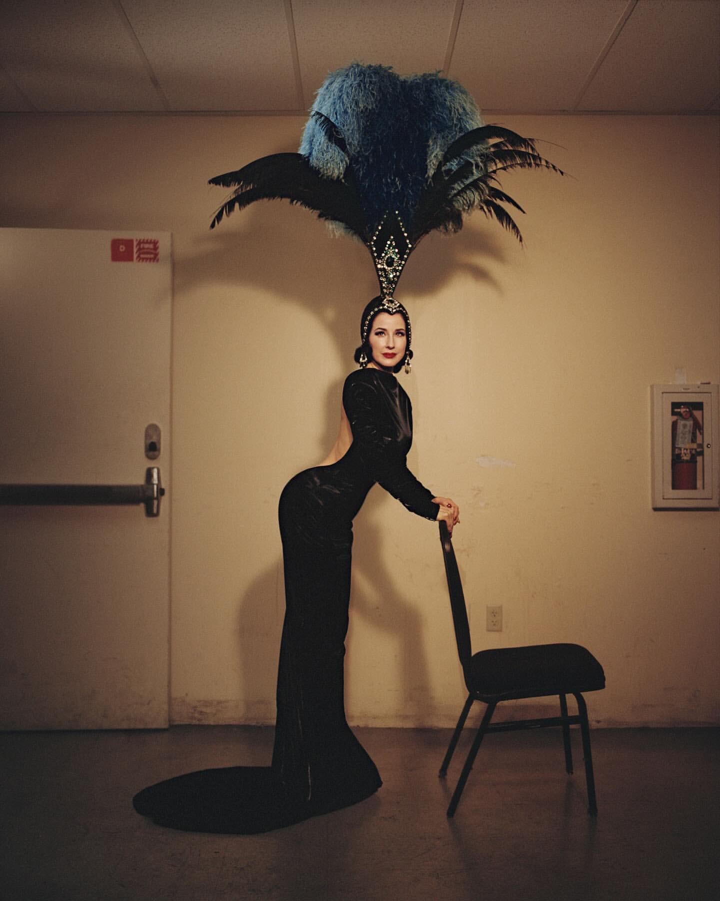 Elegant woman in black gown and elaborate feather headpiece leaning on a chair in an indoor setting with neutral colored walls.