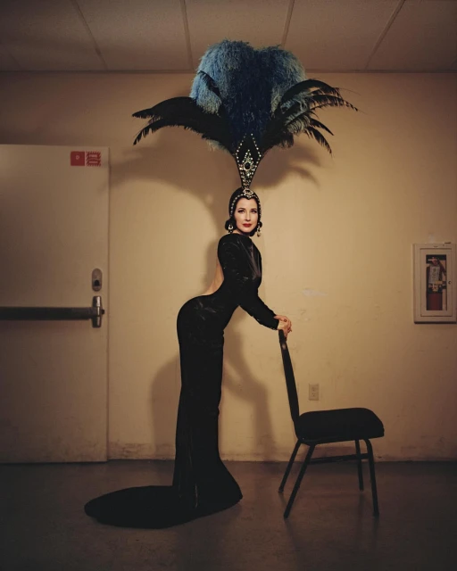 Elegant woman in black gown and elaborate feather headpiece leaning on a chair in an indoor setting with neutral colored walls.