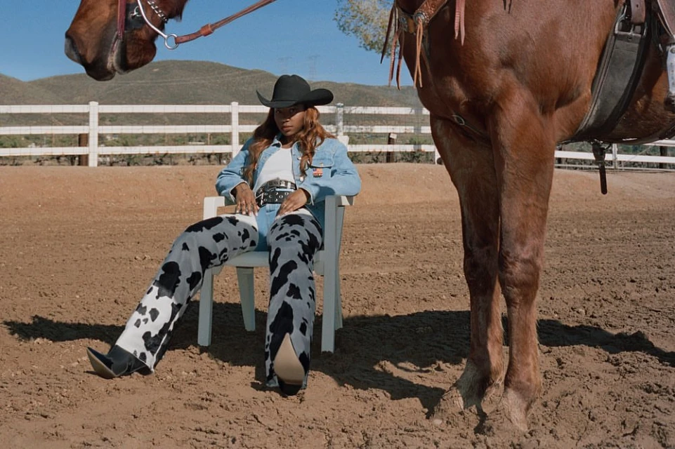 Woman in cow-print pants sits on a white chair next to a horse on a sandy lot under a blue sky with a white fence in the background.
