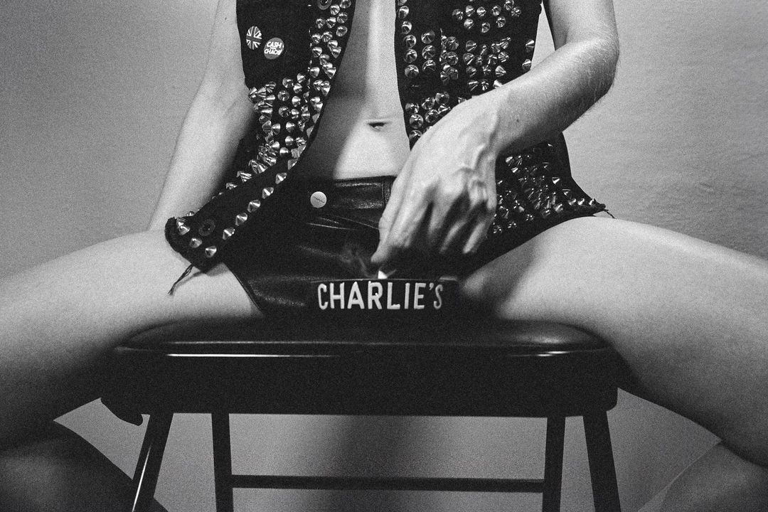 Monochrome shot of a partially visible person in studded vest and shorts, seated on a stool with an ashtray reading 'Charlie's'.