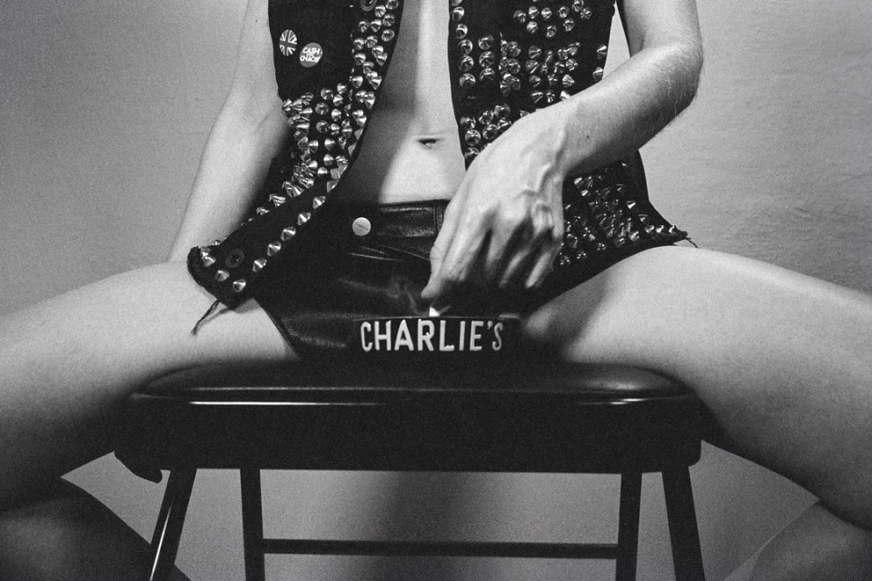 Monochrome shot of a partially visible person in studded vest and shorts, seated on a stool with an ashtray reading 'Charlie's'.