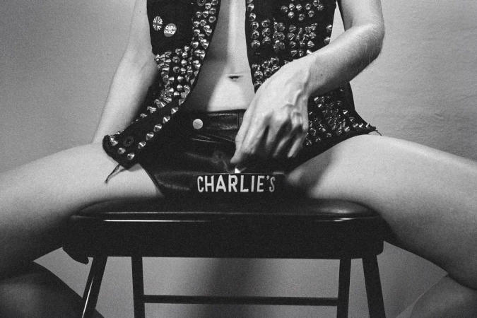 Monochrome shot of a partially visible person in studded vest and shorts, seated on a stool with an ashtray reading 'Charlie's'.