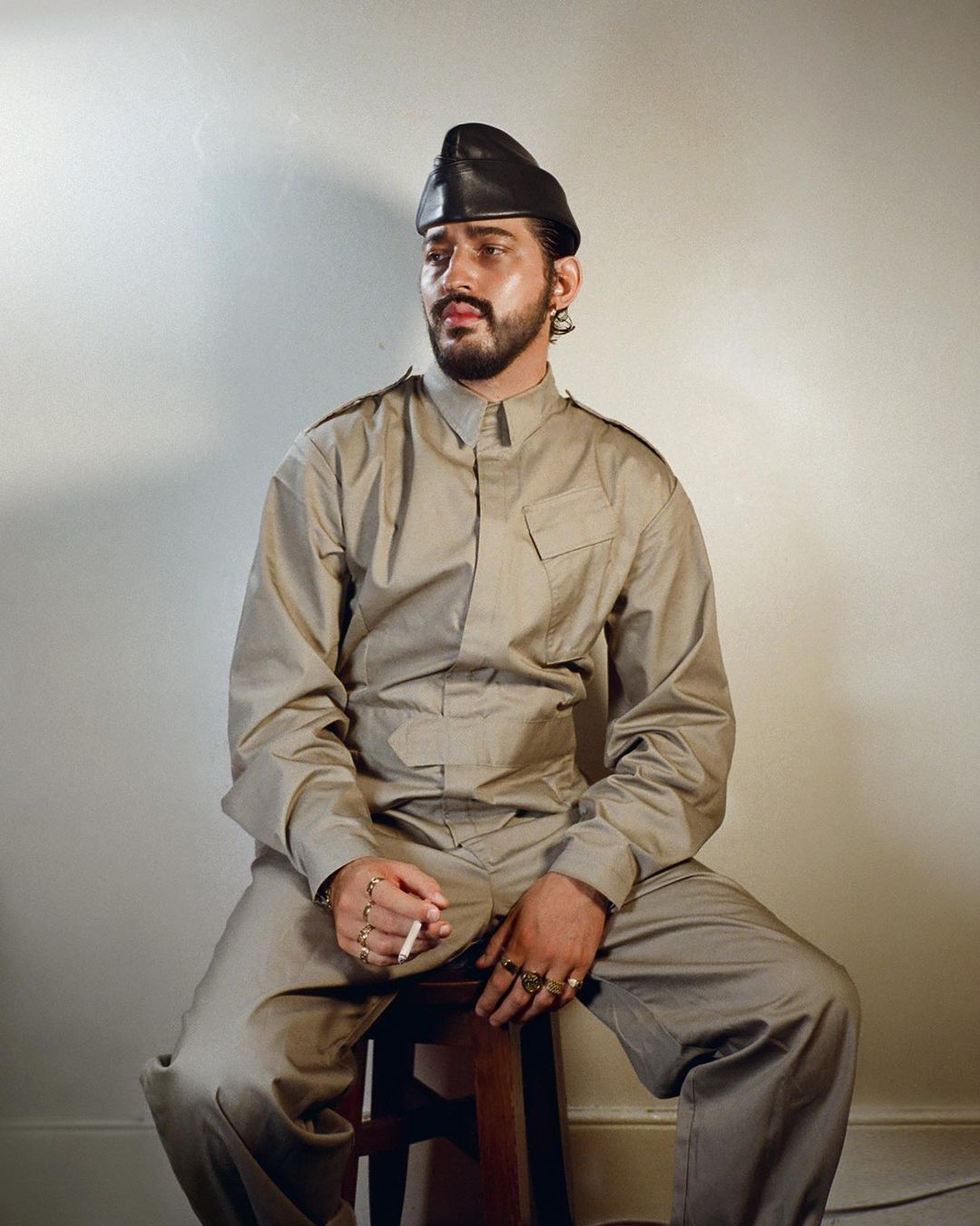 Bearded adult male in tan jumpsuit and black beret sits on wood stool holding lit cigarette in studio, looking off to the side.