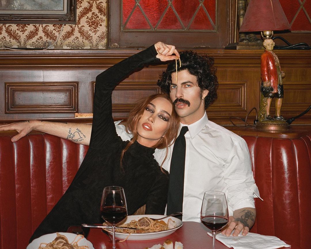 Woman and man in restaurant setting with spaghetti. Woman in black dress, man in white shirt and tie. Table with wine glasses.