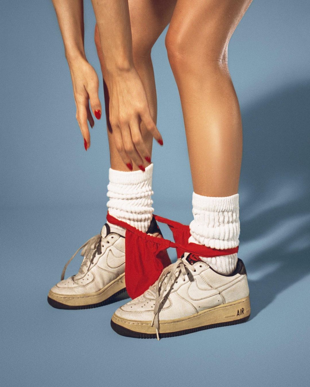 Close-up studio shot of a woman's legs and hands wearing white socks and sneakers with a red garment in front of a blue backdrop.