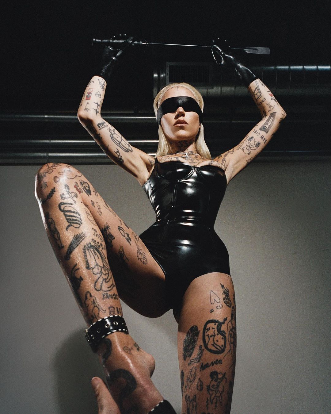 Tattooed woman wearing a black bodysuit and blindfold poses with a riding crop in a studio, shot from a low angle.