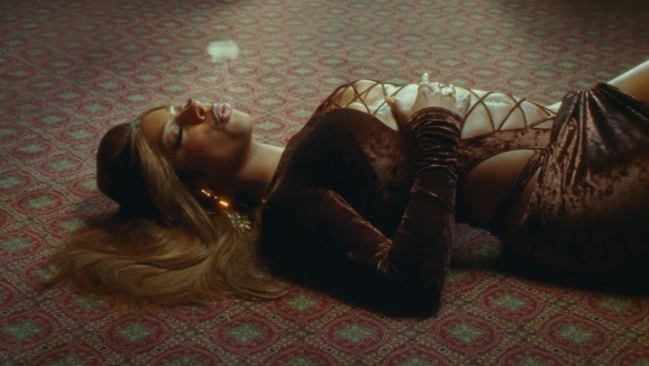 Woman with long brown hair lies on her side in a velvet brown outfit with smoke rising in an indoor environment with carpeted flooring.