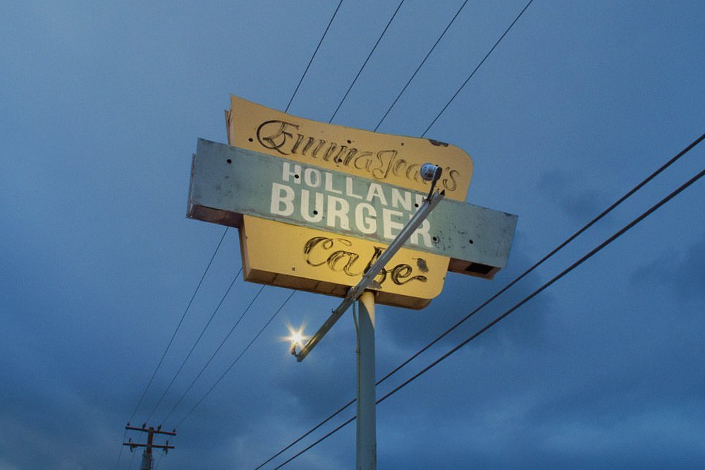 Vintage roadside sign for Emma Jeans Holland Burger Cafe against a blue dusk sky, featuring faded yellow and blue paint and power lines.