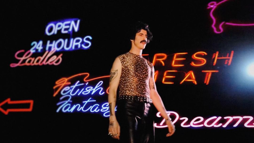 Person in leopard print top and black pants stands in front of neon signs for open hours, ladies, fresh meat, and dreams in a dark setting.