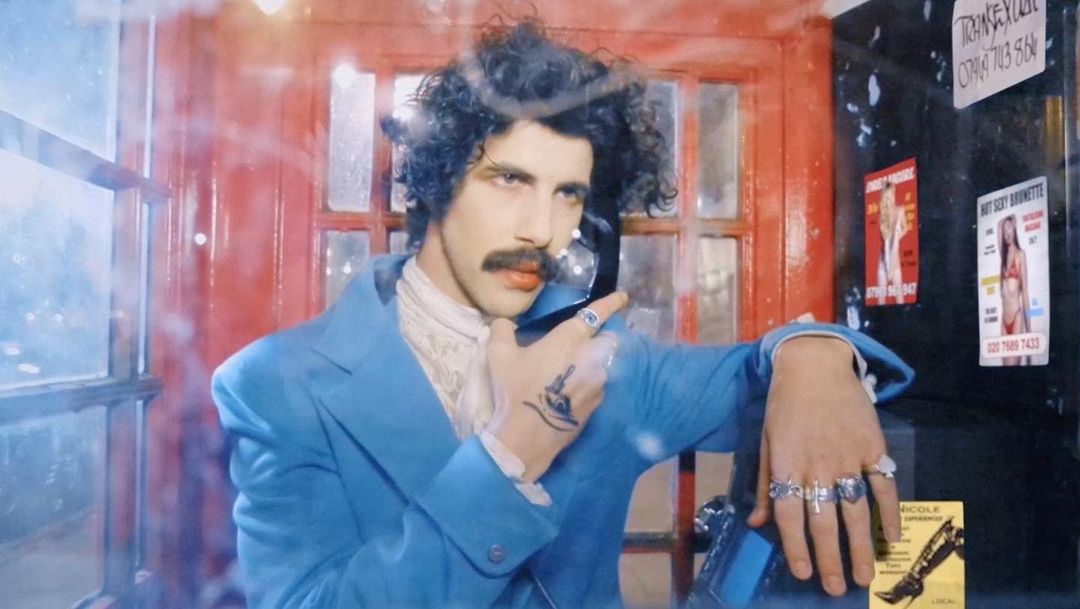 Man in a blue suit using a phone in a red phone booth with advertisements; curly hair, mustache, rings and a tattoo on his hand.