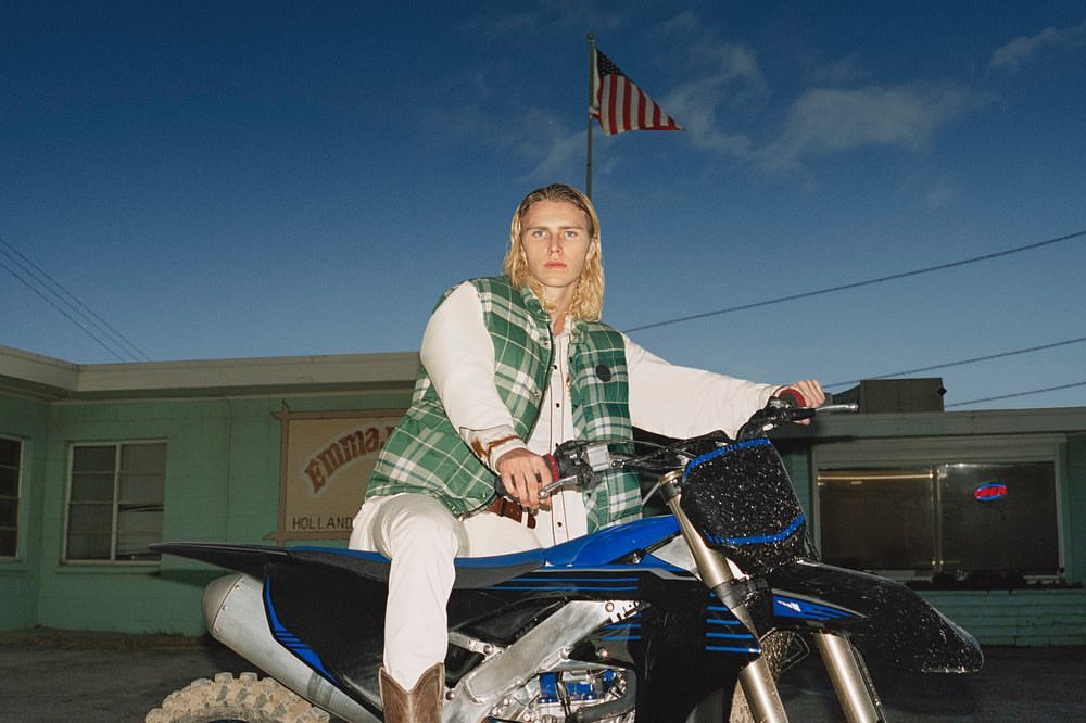 A blonde male fashion model poses on a blue dirt bike in front of Emma’s Holland restaurant with the American flag waving overhead.